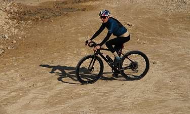 Eine Fahrerin in blauem Trikot, schwarzer Radhose und buntem Helm fährt auf einem schwarzen Gravelbike über einen hellen, trockenen Schotterweg. 