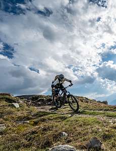Mountain biker rides down a mountain trail beneath a dramatic cloudy sky with sunbeams breaking through, with a vast alpine valley in the background