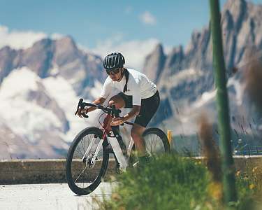 Radfahrerin auf einem Gravelbike fährt auf einer Bergstrasse vor beeindruckender Alpenkulisse mit schneebedeckten Gipfeln im Hintergrund.