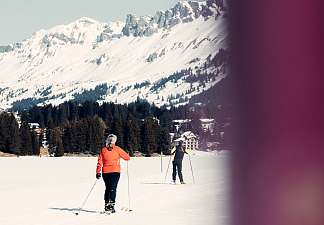 Zwei Frauen auf Langlaufskis in der verschneiten Winterlandschaft der Lenzerheide, im Hintergrund Nadelbäume und Bergpanorama.